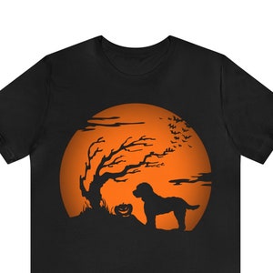 May include: A black short-sleeve t-shirt featuring a graphic print of a dog standing next to a jack-o'-lantern under a large orange full moon. The scene includes a silhouette of a gnarled tree and a flock of bats flying across the moon.