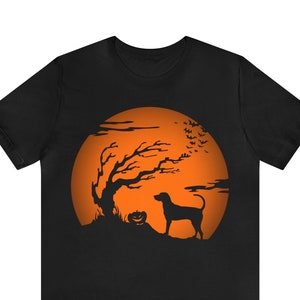 May include: A black short-sleeved t-shirt featuring a large orange circular graphic. The design depicts a black silhouette of a dog standing next to a jack-o'-lantern and a bare tree, with a flock of small bats flying against the orange moon background.