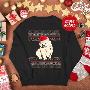 Pode incluir: Camisola de Natal preta com um cão branco a usar um chapéu de Pai Natal vermelho sobre um fundo estampado vermelho e verde. O texto "mais cores" está num círculo vermelho.