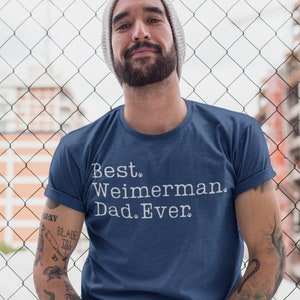 以下が含まれることがあります： 白文字で「Best. Weimaraner. Dad. Ever.」と書かれたネイビーブルーのTシャツ