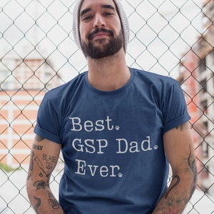 May include: Navy blue t-shirt with the text "Best GSP Dad Ever" in white. The shirt has rolled-up sleeves. The person in the image is wearing a gray beanie and has tattoos on their arm.