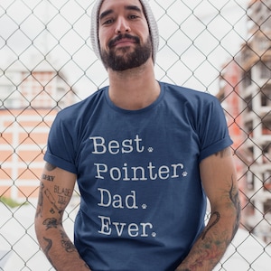 Puede incluir: Un hombre con una camiseta azul con el texto "Best Pointer Dad Ever" en blanco con huellas de patas. Está de pie frente a una valla de cadena.
