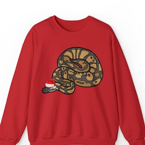 Peut inclure: Sweat-shirt rouge avec une image de dessin animé d'un serpent portant un chapeau de Père Noël.