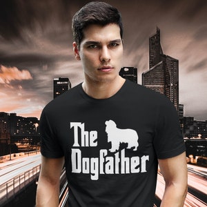 Pode incluir: Camiseta preta com a frase "The Dogfather" e uma silhueta branca de cachorro. A pessoa está usando jeans azuis, em um fundo de horizonte da cidade.