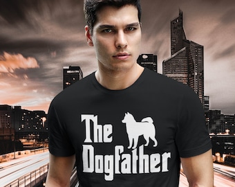 May include: Black t-shirt with the text "The Dogfather" and a white silhouette of a dog. The shirt is worn by a person in blue jeans. The background features a cityscape with blurred lights.