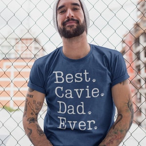 Pode incluir: Um homem vestindo uma camiseta azul marinho com o texto "Best Cavie Dad Ever" em branco com pegadas de patas. O homem tem barba e está usando um gorro branco.