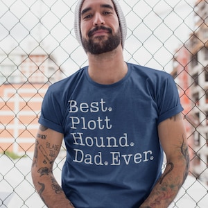 Può includere: Un uomo indossa una maglietta blu con la scritta "Best Plott Hound Dad Ever" stampata in bianco. Il testo è circondato da impronte di zampe.