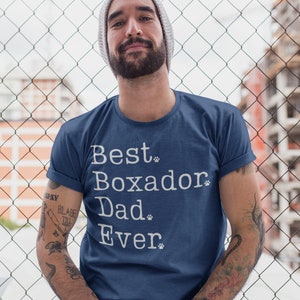 May include: A man wearing a blue t-shirt with the text "Best Boxador Dad Ever" in white with paw prints. He is standing in front of a chain link fence.