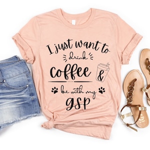 May include: A peach colored t-shirt with the text "I just want to drink coffee & be with my GSP" in black. The text is surrounded by paw prints and a coffee cup graphic.