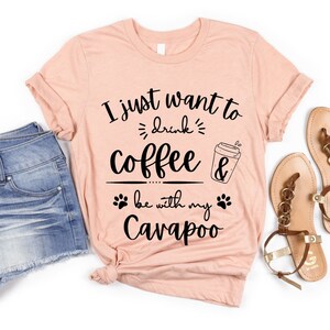 Könnte beinhalten: Ein pinkfarbenes T-Shirt mit dem Text "I just want to drink coffee & be with my Cavapoo" in Schwarz. Das Shirt wird mit Jeans-Shorts und braunen Sandalen kombiniert.
