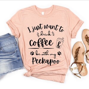 Può includere: Una maglietta color pesca con il testo "I just want to drink coffee & be with my Peekaboo" in nero. La maglietta è indossata con pantaloncini di jeans e sandali marroni.
