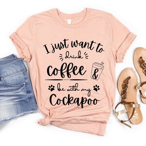 May include: A peach-colored t-shirt with the text "I just want to drink coffee & be with my Cockapoo" in black. The text is in a handwritten font and includes a coffee cup and paw prints.