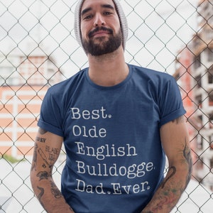 May include: A man wearing a blue t-shirt with white text that reads "Best. Olde English Bulldogge. Dad. Ever." He is wearing a beanie and has tattoos on his arms.
