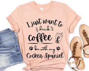 Puede incluir: Una camiseta de color melocotón con el texto "I just want to drink coffee & be with my Cocker Spaniel" en negro. La camiseta se combina con pantalones cortos de mezclilla y sandalias marrones.