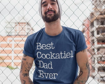 May include: A man wearing a blue t-shirt with the text "Best Cockatiel Dad Ever" in white. He is standing in front of a chain link fence.