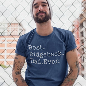 Puede incluir: Un hombre con una camiseta azul con el texto "Best Ridgeback Dad Ever" en blanco. La camiseta tiene huellas de patas al lado de las palabras "Ridgeback" y "Ever".
