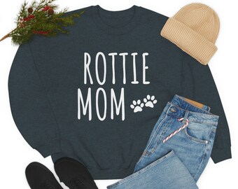 May include: A dark blue sweatshirt with the text "Rottie Mom" in white with two white paw prints. The sweatshirt is laid out on a white background with a pair of black sneakers, blue jeans, and a beige beanie.