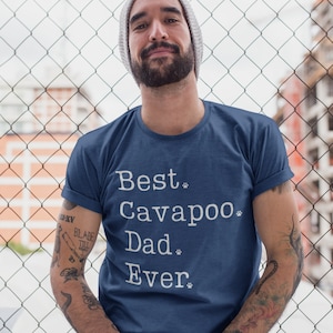 Könnte beinhalten: Marineblaues T-Shirt mit dem weißen Text "Best Cavapoo Dad Ever". Das Shirt hat hochgekrempelte Ärmel. Die Person auf dem Bild trägt eine graue Mütze.
