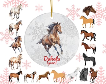 May include: A round ceramic Christmas ornament with a running brown horse illustration. The ornament has a gold string for hanging and features the text "Dakota [year]" in red. Surrounding the ornament are images of various horse breeds and pink snowflakes.