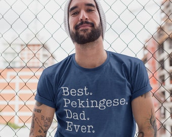 Könnte beinhalten: Ein Mann trägt ein marineblaues T-Shirt mit weißem Text, der "Best Pekingese Dad Ever." lautet. Der Text ist von kleinen Pfotenabdrücken umgeben. Der Mann hat einen Bart und trägt eine Mütze.