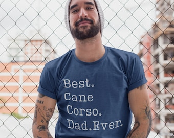 Può includere: Un uomo indossa una maglietta blu con la scritta "Best Cane Corso Dad Ever" stampata in bianco. La maglietta ha delle impronte di zampa intorno al testo. L'uomo ha la barba e indossa un berretto.