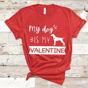 Puede incluir: Camiseta roja con la frase "My dog is my Valentine" en blanco. Un diseño con la silueta blanca de un perro con un corazón. La camiseta está anudada en la parte inferior.
