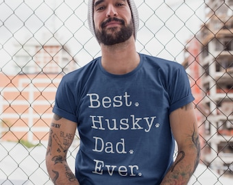 May include: A man wearing a blue t-shirt with the text "Best Husky Dad Ever" printed in white. He is standing in front of a chain-link fence.