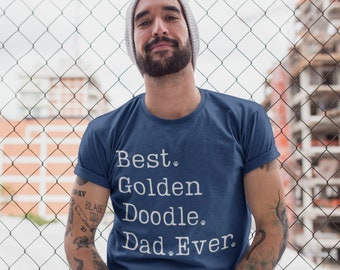 May include: A man wearing a blue t-shirt with the text "Best Golden Doodle Dad Ever" printed on it. He is standing in front of a chain link fence.