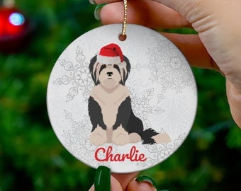 May include: A round, white Christmas ornament featuring a black and white dog wearing a red Santa hat. The dog's name, "Charlie," is written in red script below the dog. The ornament has a gold string for hanging and a snowflake background.