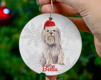May include: A round, white Christmas ornament featuring a watercolor illustration of a dog wearing a Santa hat. The dog's name, "Bella," is written in red script below the image. The ornament has a gold string for hanging.