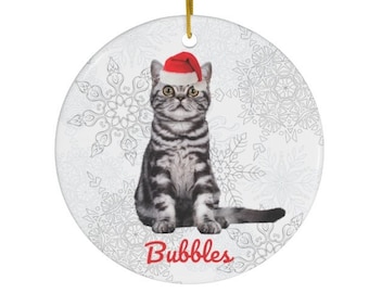May include: A round ceramic Christmas ornament featuring a gray tabby cat wearing a red Santa hat. The cat is sitting on a white background with a snowflake pattern. The ornament has the word "Bubbles" written in red at the bottom.