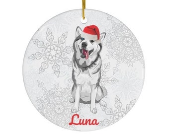 May include: A round, white ceramic ornament featuring a smiling husky wearing a Santa hat. The ornament has a gold string for hanging and is decorated with snowflakes. The name "Luna" is written in red script.