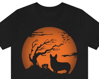 Op de afbeelding: Een zwart t-shirt met korte mouwen en een grafische print van het silhouet van een Corgi-hond die naast een pompoenlantaarn en een kale boom staat tegen een grote, ronde oranje maan met een zwerm kleine vleermuizen op de achtergrond.