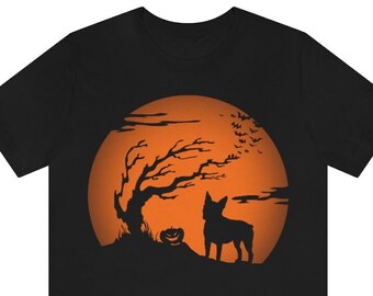 Puede incluir: Una camiseta negra de manga corta con un estampado gráfico de un perro de pie sobre una colina frente a una gran luna llena naranja. El diseño incluye la silueta de un árbol sin hojas, una pequeña calabaza y una bandada de murciélagos.