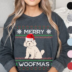 Puede incluir: Una sudadera gris oscuro con un copo de nieve blanco y el texto "Merry Woofmas" en blanco. Un perro blanco con un gorro de Papá Noel rojo está en el centro del diseño.