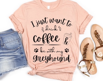 以下が含まれることがあります： ピンクのTシャツに、黒い文字で「I just want to drink coffee & be with my Greyhound」と書かれています。Tシャツにはコーヒーカップと足跡が描かれています。