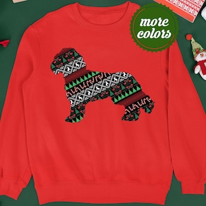 Op de afbeelding: Rode kersttrui met een silhouet van een hond die een feestelijke trui met patroon draagt. De trui heeft een "meer kleuren" label.