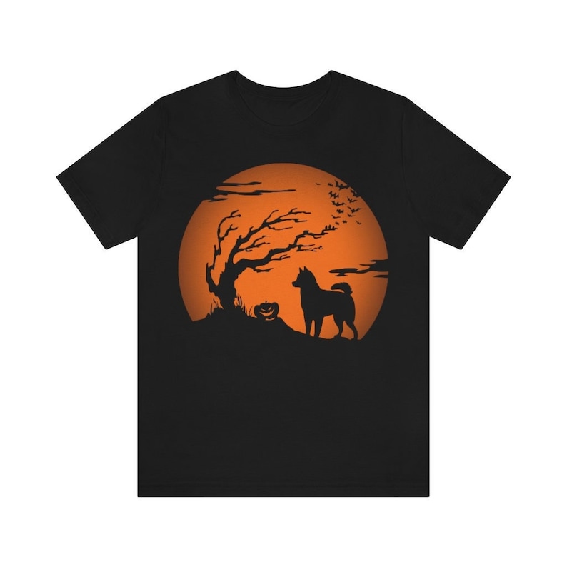 May include: A black short-sleeve t-shirt featuring a graphic print of a dog standing next to a jack-o'-lantern and a bare tree against a large orange full moon. The scene includes silhouettes of flying bats in the upper right corner.