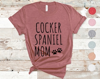 Puede incluir: Una camiseta color marrón rojizo con el texto "Cocker Spaniel Mom" en letras negras y dos huellas de patas negras.