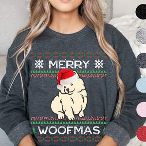 Pode incluir: Camisola cinza escuro com um design festivo. A camisola apresenta o texto "MERRY WOOFMAS" com um cão de desenho animado usando um chapéu de Papai Noel. O design inclui flocos de neve, árvores de Natal e detalhes de bengalas doces. Disponível em várias cores.