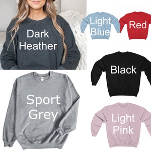 May include: A selection of crewneck sweatshirts in various colors, including dark heather, light blue, red, black, sport grey, and light pink. Each sweatshirt has the color name printed in white on the front. The sweatshirts are made of a soft material.