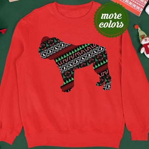 May include: Red Christmas sweater with a silhouette of a bulldog wearing a festive patterned sweater. The sweater has a green, red, and white pattern with candy canes, reindeer, and Christmas trees. The text "more colors" is on a green circle in the upper right corner of the image.