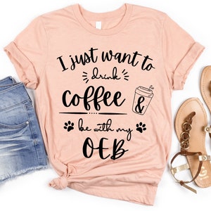 May include: A light peach-colored t-shirt with the text "I just want to drink coffee & be with my OEB" in black. The shirt features a coffee cup graphic and paw prints. The shirt is paired with denim shorts and sandals.