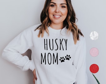 Op de afbeelding: Witte trui met de tekst "Husky Mom" en twee poot afdrukken in zwart.