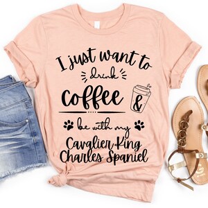 Puede incluir: Una camiseta de color melocotón con el texto "I just want to drink coffee & be with my Cavalier King Charles Spaniel" impreso en negro.