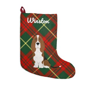 Puede incluir: Calcetín navideño con un estampado de cuadros rojos, verdes y amarillos. El calcetín presenta una ilustración de un perro basset y el nombre "Winston" en escritura blanca. Un lazo rojo en la parte superior.