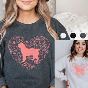 May include: A dark gray sweatshirt with a pink heart design featuring a silhouette of a dog inside. The heart is surrounded by a floral pattern. The sweatshirt is shown on a model.