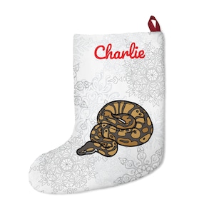 Peut inclure: Chaussette de Noël blanche avec une boucle de suspension rouge et le nom "Charlie" en script rouge. La chaussette présente une illustration détaillée d'un python royal dans des tons de marron et de beige. Le fond a un motif de flocon de neige subtil.