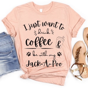 May include: A pink t-shirt with the text "I just want to drink coffee & be with my Jack-A-Poo" in black lettering. The shirt is laid out on a white surface with a pair of brown sandals.