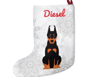Op de afbeelding: Witte kerstsok met een illustratie van een Dobermann Pinscher. De hond is zwart en bruin, met de naam "Diesel" in rood script erboven. De achtergrond heeft een sneeuwvlokkenpatroon en een rode ophanglus.